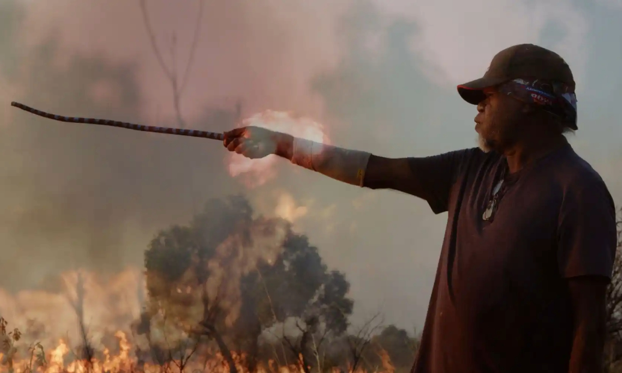 Profile of a First Nations man in a cap holding out a stick as fire rages in the background.