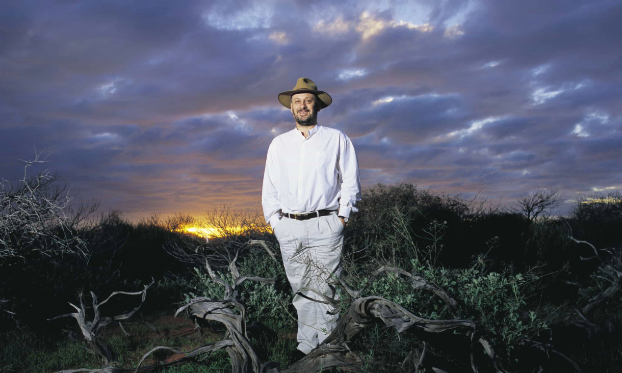 Man in white shirt and pants wearing a hat stands in a moody outdoor scene amongst bushes with a cloudy dusk sky above.