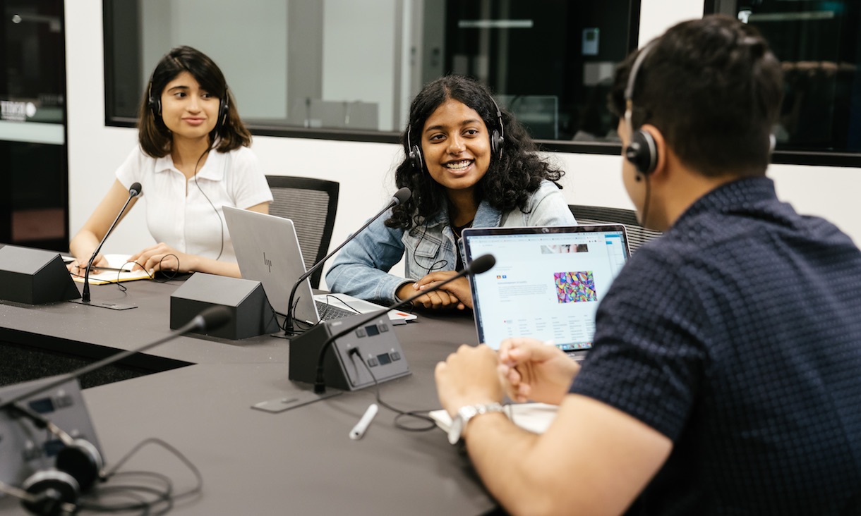 Students wearing earmuffs talk to each other in a recording studio.
